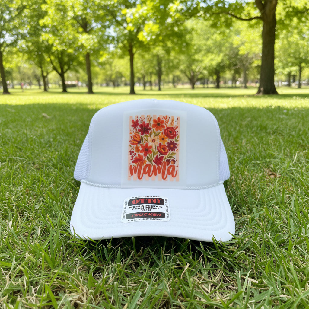 A white trucker hat with a colorful floral patch on the front that says 'mama' in orange script. The hat has a visible OTTO brand sticker on the brim and is photographed on a wooden surface.