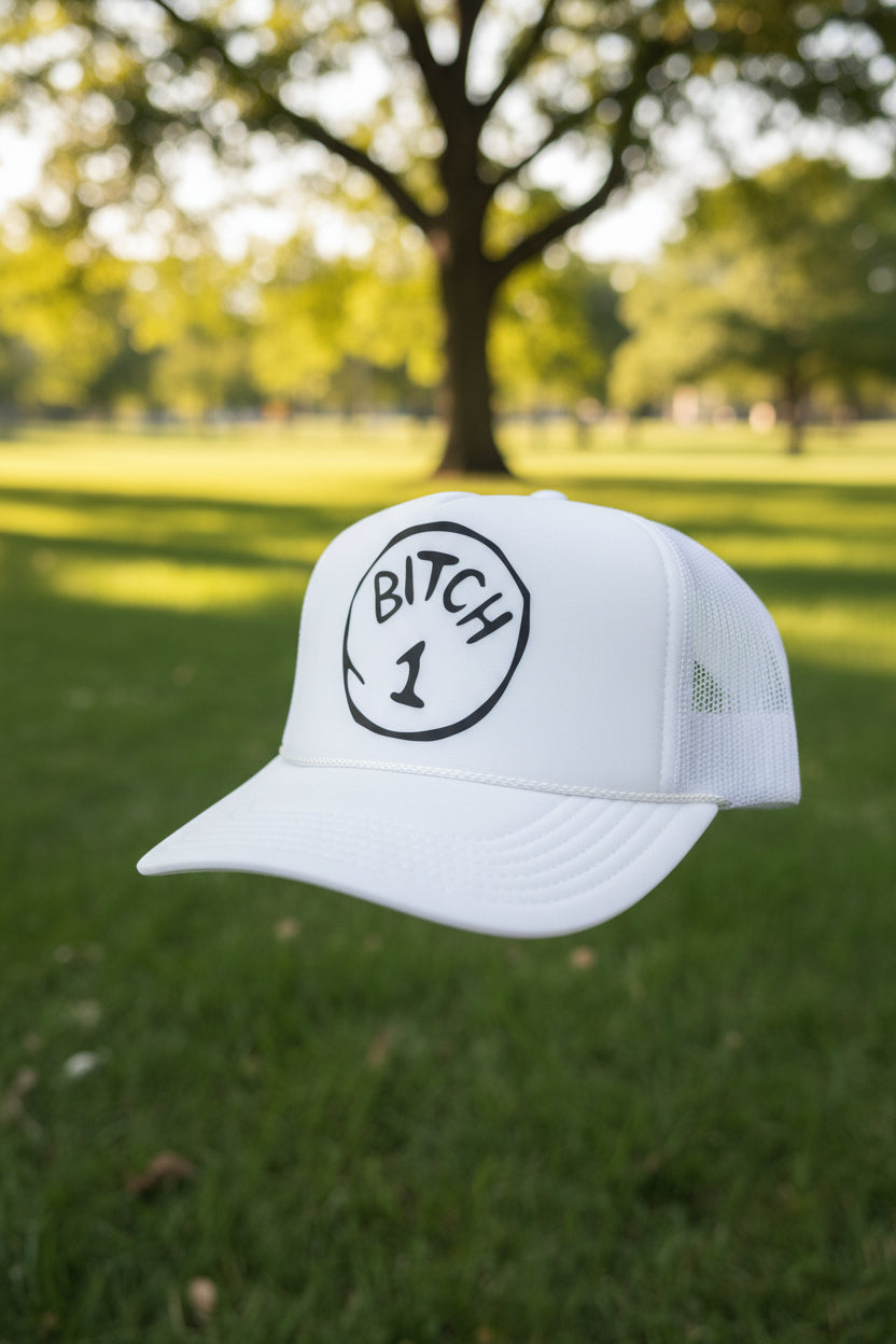 A white trucker hat with a black circular graphic on the front that reads 'BITCH 1' .