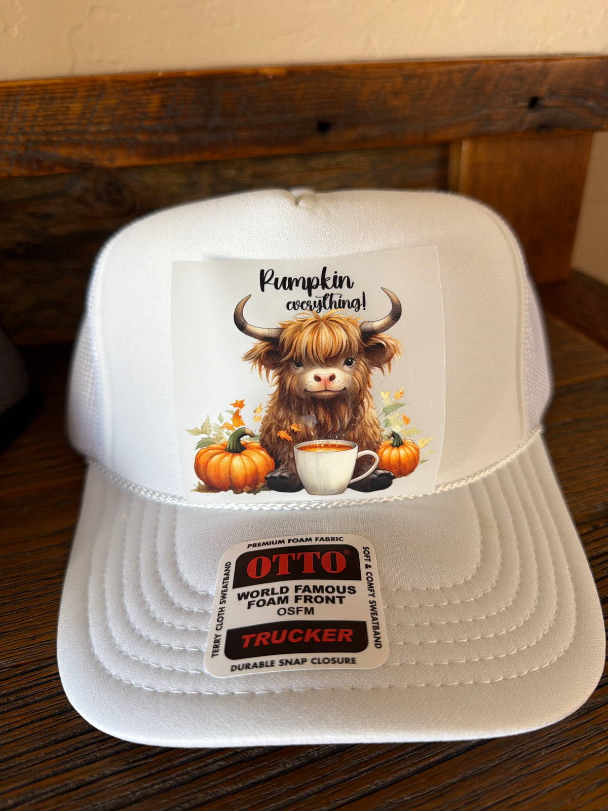 A white trucker hat with a custom graphic of a shaggy Highland cow surrounded by pumpkins and a coffee cup, sitting on a dark wooden surface. The hat features a visible 'OTTO' brand sticker on the brim.