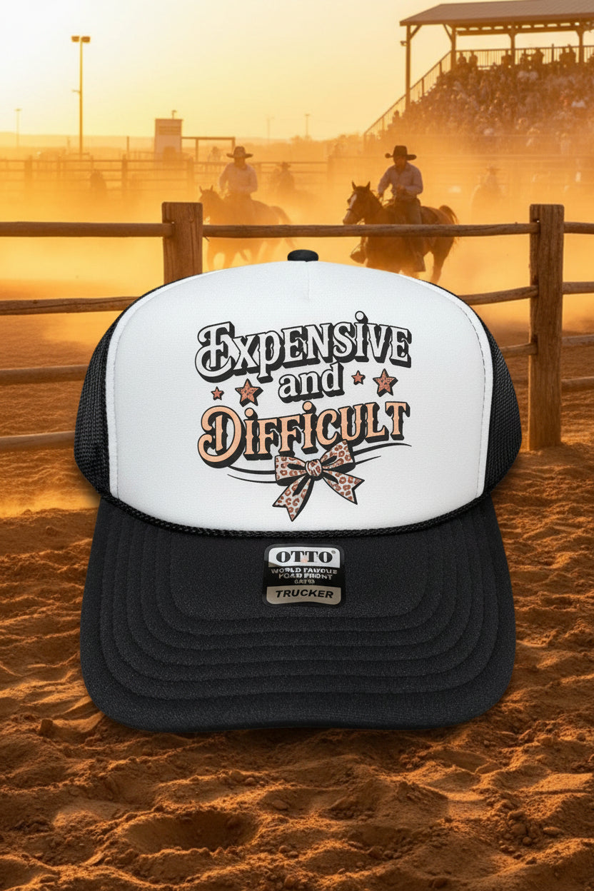 A black and white trucker hat with a white front panel and black mesh back. The front panel features the text 'Expensive and Difficult' in stylized lettering with a leopard-print bow graphic.
