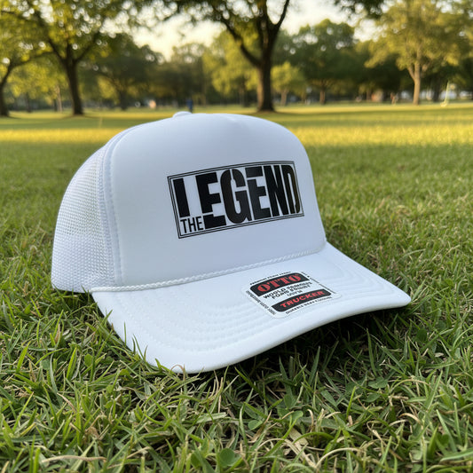 A white trucker hat with a black 'LEGEND THE' graphic on the front panel, resting on a wooden surface. The brim features a white sticker with 'OTTO TRUCKER' branding.