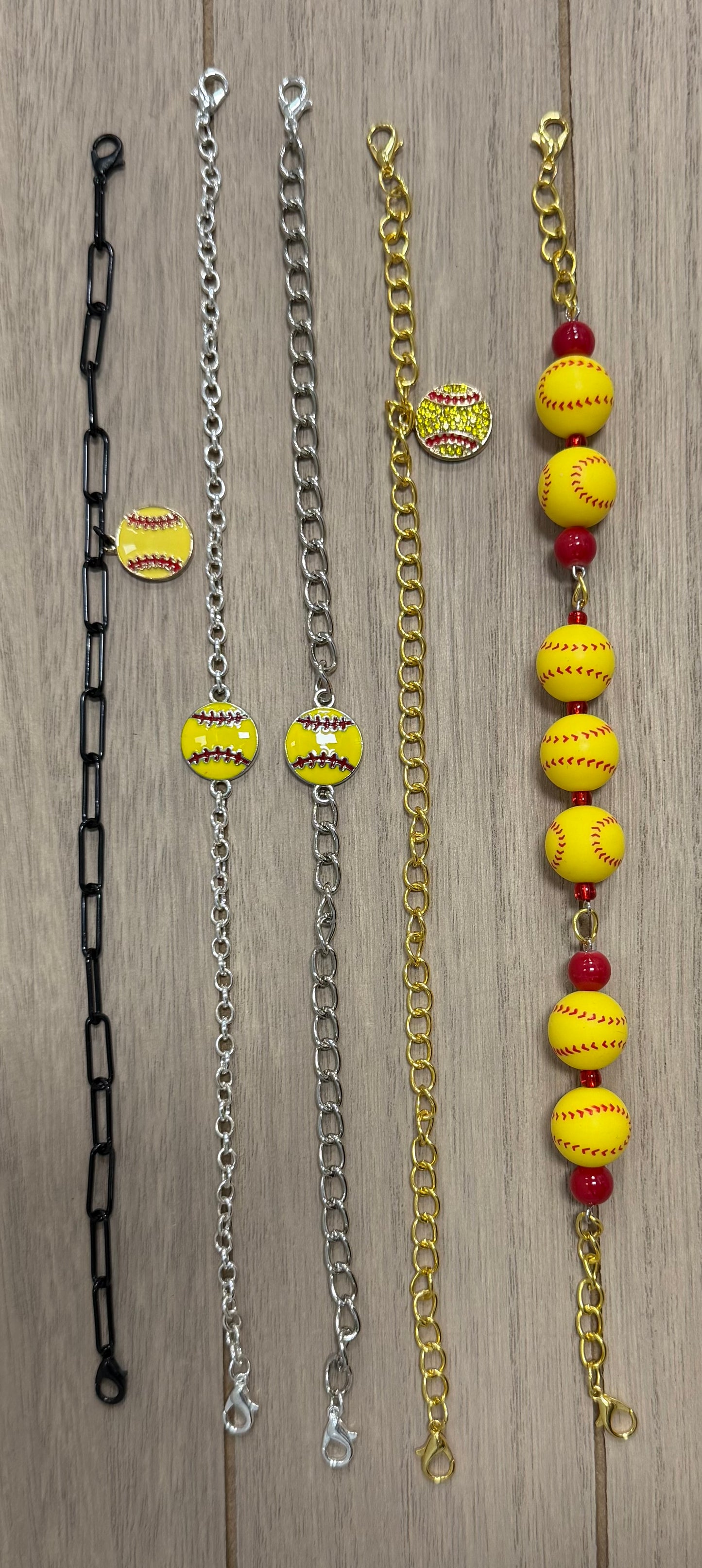  Softball hat chains laid out on a light wood background.  Featuring black, silver, gold, and gold-with-red-bead chains, each embellished with small yellow softball charms and larger yellow softball beads with red spacer beads.