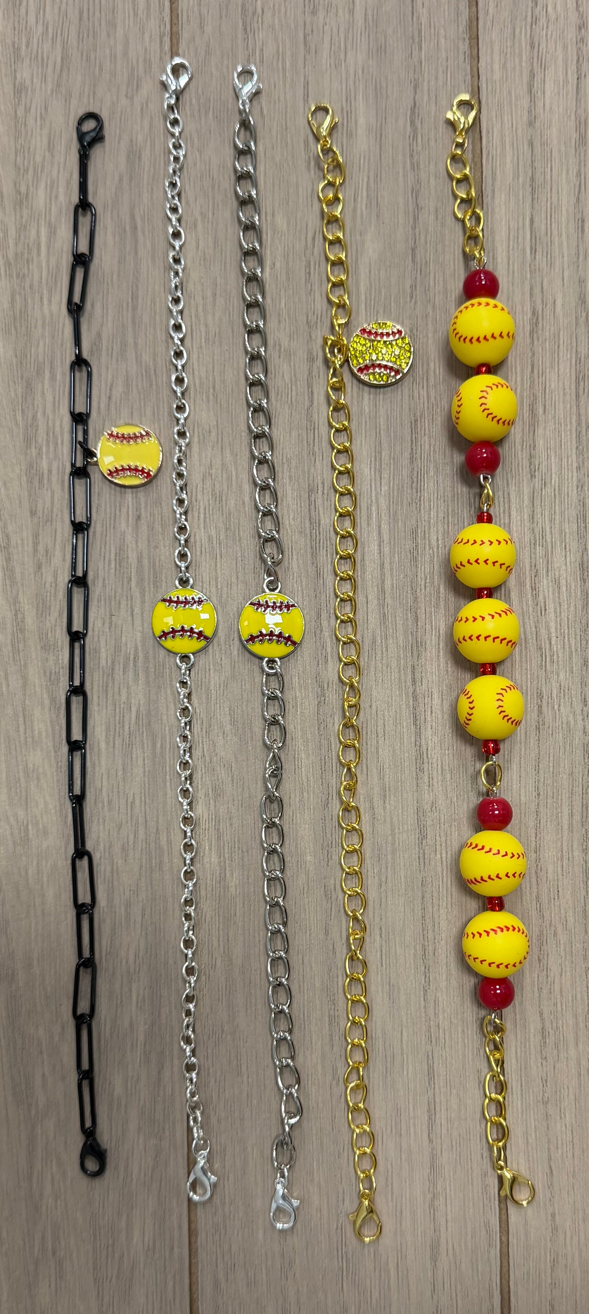  Softball hat chains laid out on a light wood background.  Featuring black, silver, gold, and gold-with-red-bead chains, each embellished with small yellow softball charms and larger yellow softball beads with red spacer beads.
