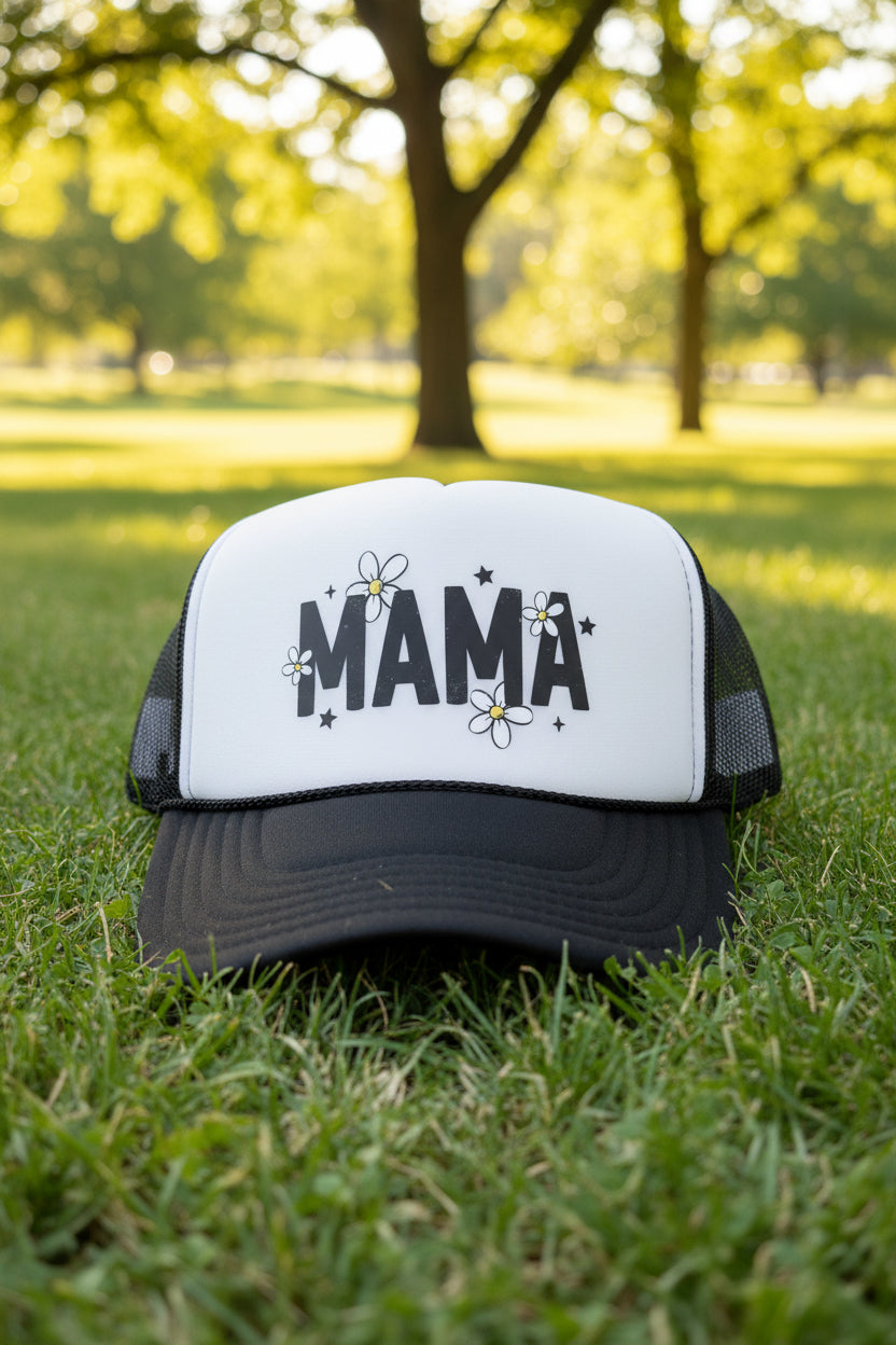 A black and white trucker hat with 'MAMA' and daisy graphics on the front, displayed on a wooden table.