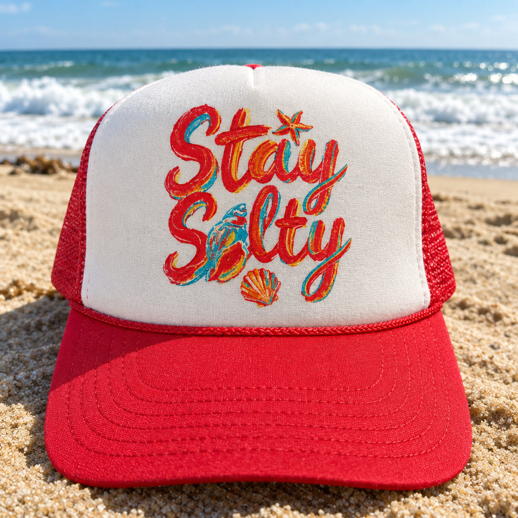 A red and white trucker hat with 'Stay Salty' written in orange and blue script on the front, resting on a wooden surface. The brim has a visible Otto Trucker patch.