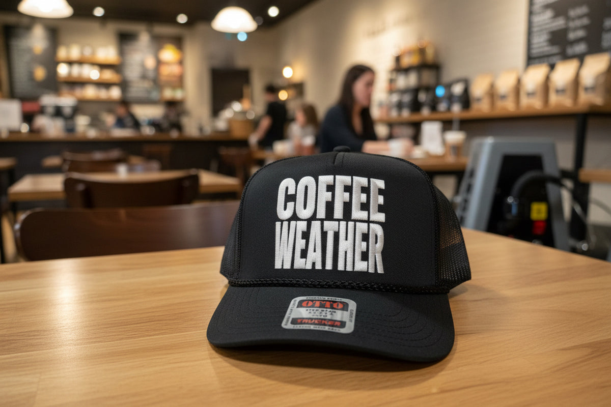 A black trucker hat with a gray rectangular patch on the front that reads 'COFFEE WEATHER' in large white letters. The brim has a visible Otto Five Star Coin Trucker sticker.