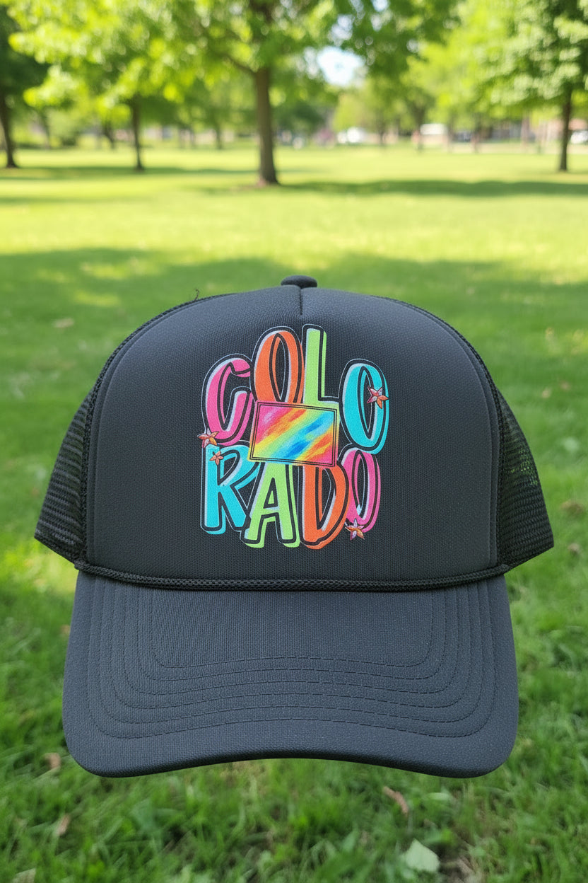 A dark gray trucker hat with a colorful 'COLORADO' graphic on the front, set against a blurred green park background.