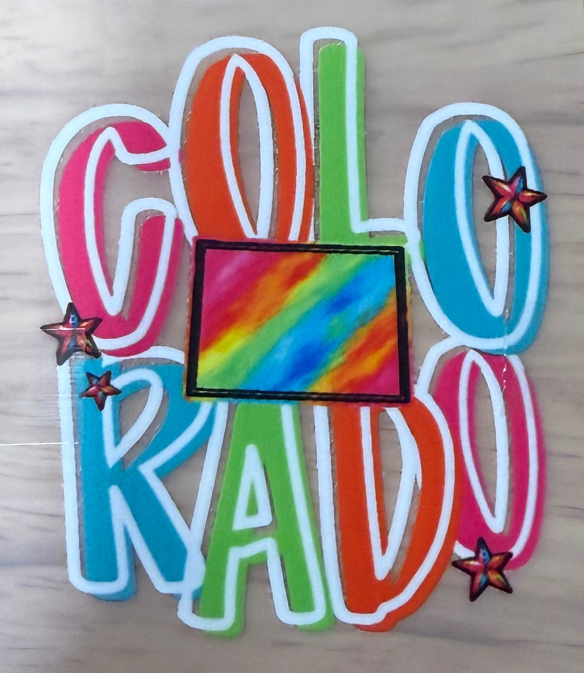 A square decal sticker featuring the word 'COLORADO' in rainbow-colored letters with a central rainbow flag and small star accents, shown on a light wood surface.
