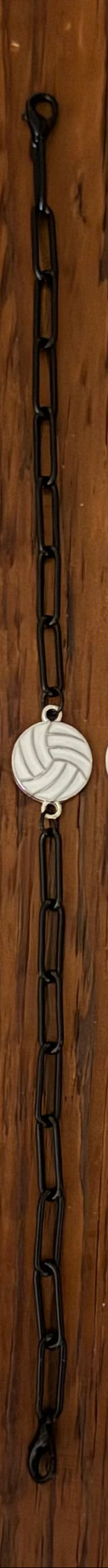 Four beaded and chain bracelets laid flat on a wooden surface, featuring black and white beads, white and silver volleyball charms, and one yellow-and-blue volleyball charm.