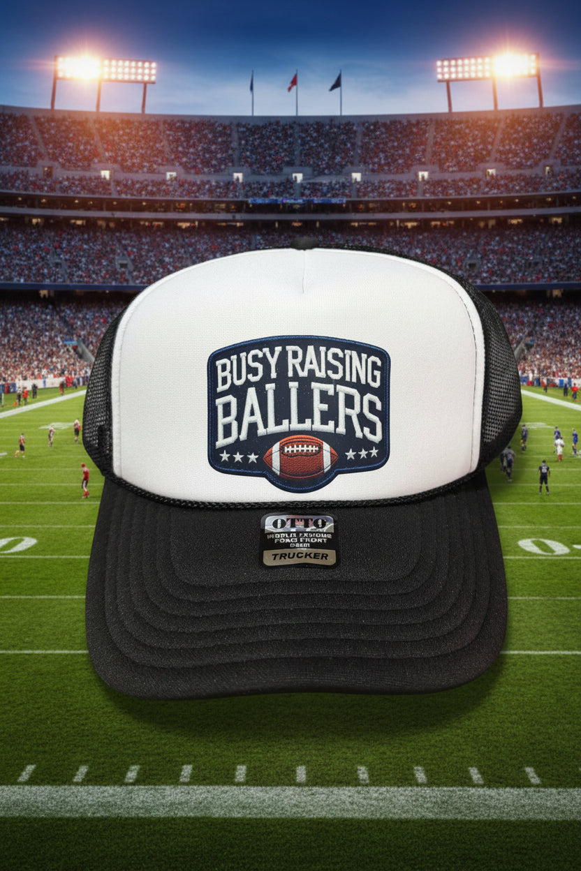 A black and white trucker hat with a 'BUSY RAISING BALLERS' football patch on the front. The brim is black, and the mesh back is white.