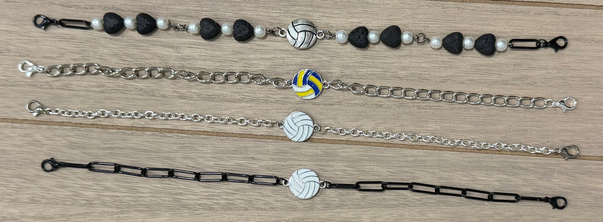 Four different volleyball-themed chain bracelets laid out horizontally on a light wood background. The bracelets feature silver and black chains, white and black beads, and a central white volleyball charm with yellow and blue accents.