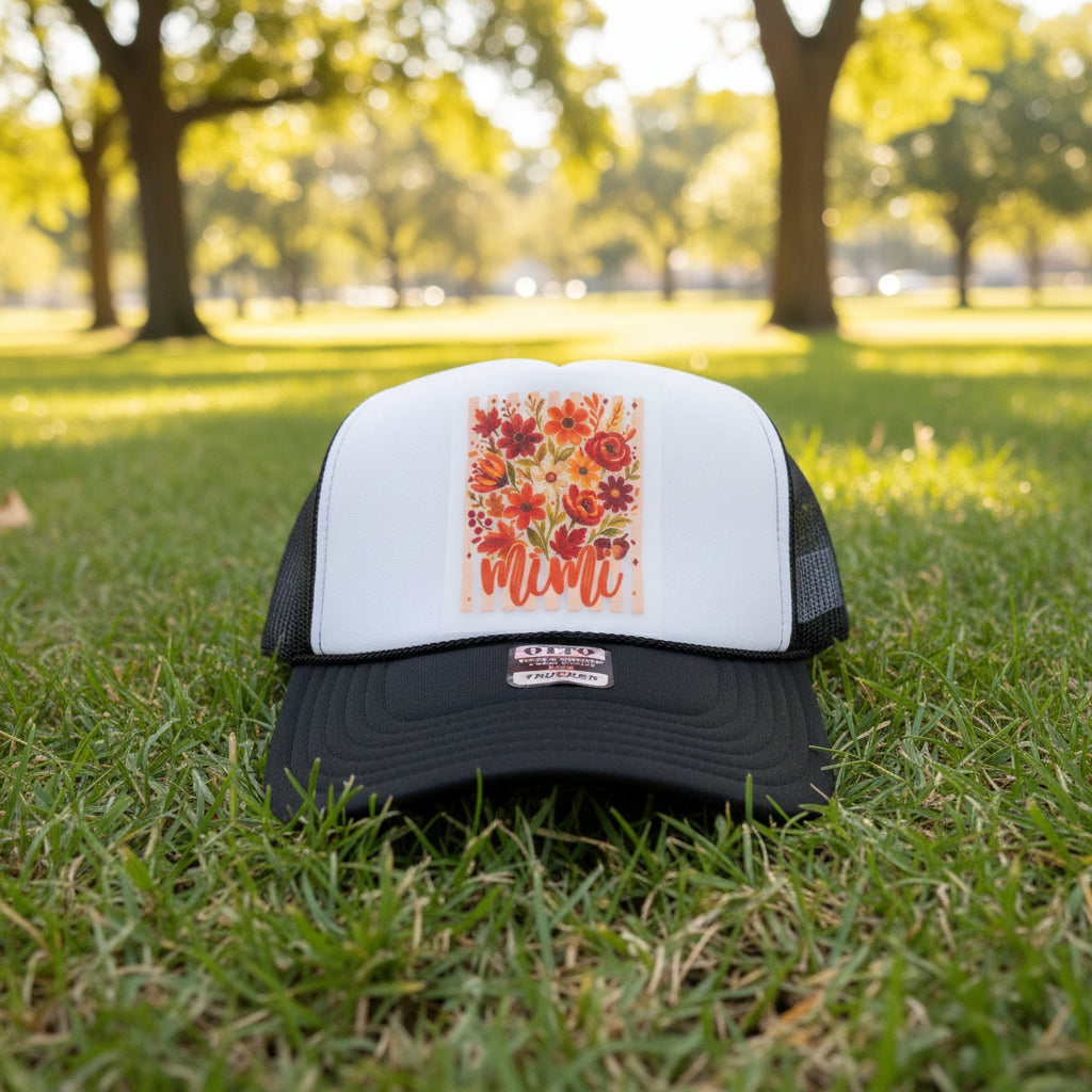 A black and white trucker hat with a colorful floral patch on the front that says 'mimi' in orange script. The hat is resting on a rustic wooden shelf, with another blue and white hat partially visible on the right.