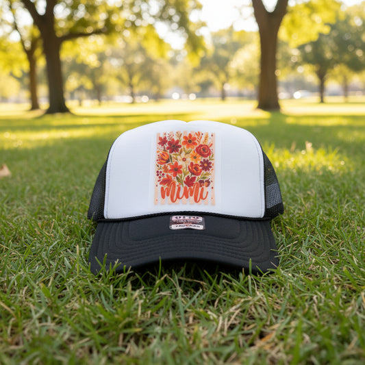 A black and white trucker hat with a colorful floral patch on the front that says 'mimi' in orange script. The hat is resting on a rustic wooden shelf, with another blue and white hat partially visible on the right.