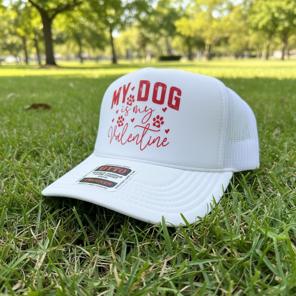 A white trucker hat with red text reading 'MY DOG is my Valentine' and paw prints, photographed on a wooden surface with a wooden chair in the background.