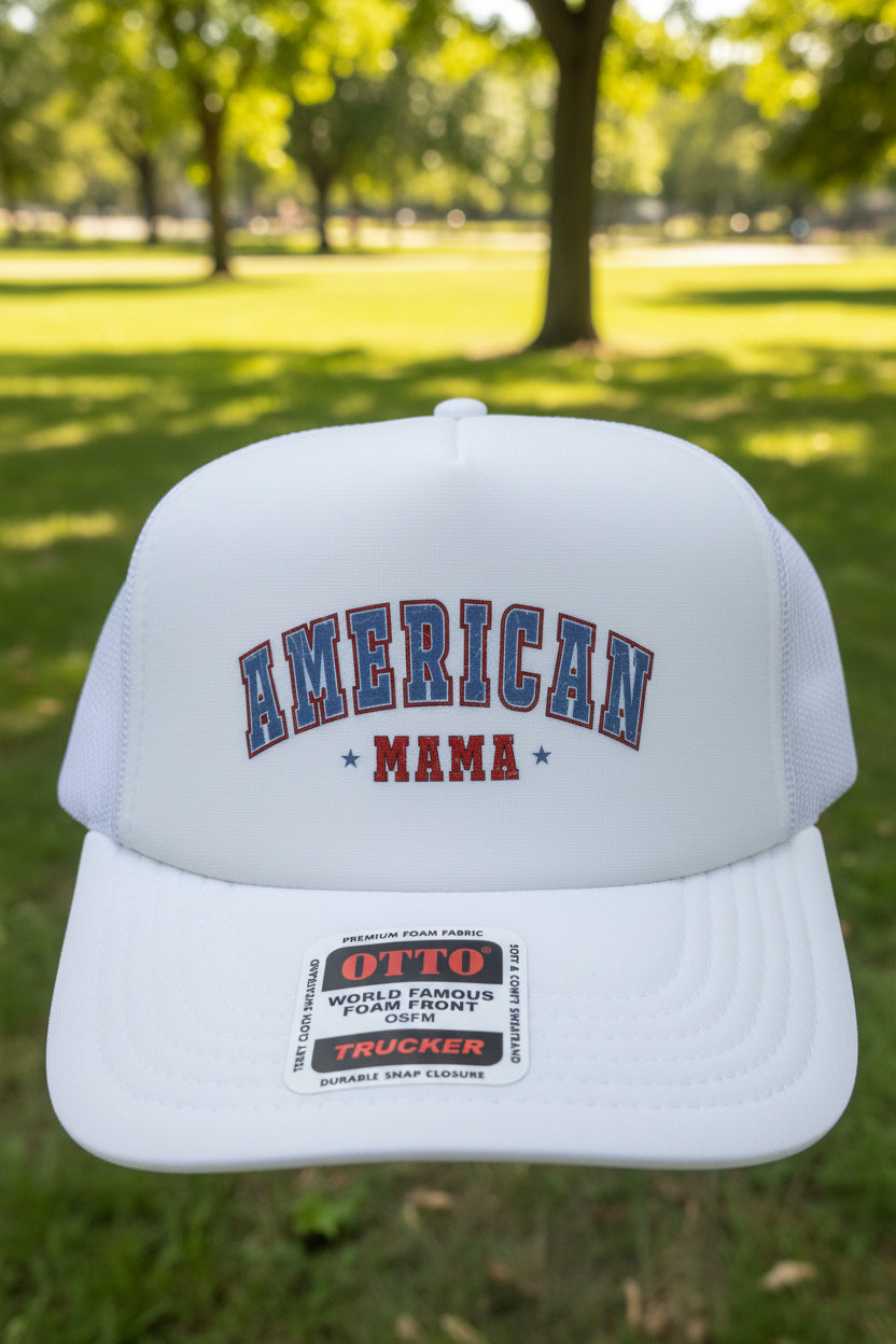 A white trucker hat with 'AMERICAN MAMA' printed on the front in blue and red, resting on a wooden surface.
