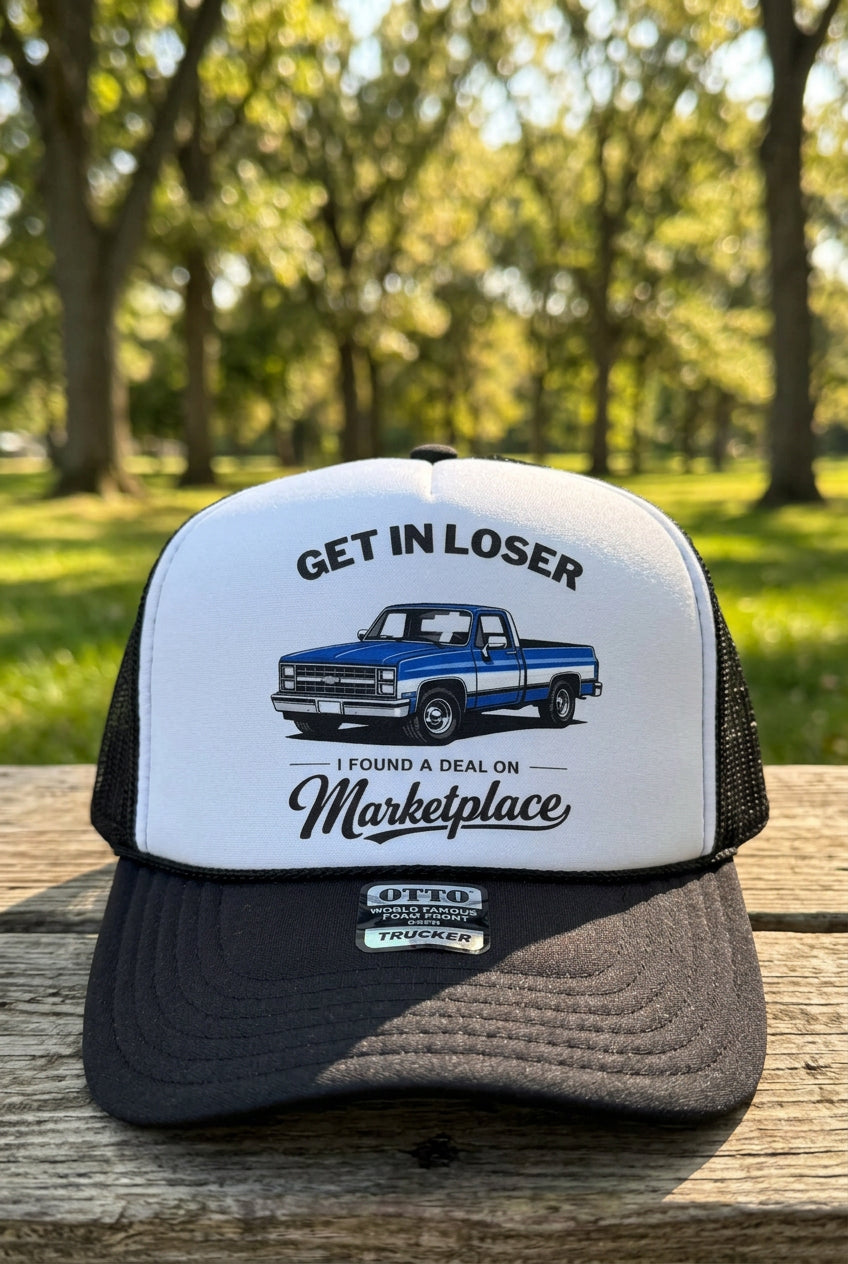 A black and white trucker hat with a graphic print of a blue pickup truck and the text 'GET IN LOSER I FOUND A DEAL ON Marketplace'. The hat is resting on a light wood surface.