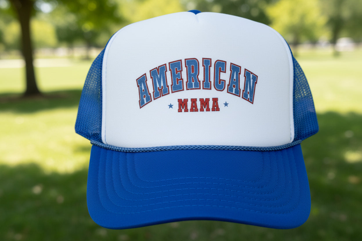 A blue and white trucker hat with 'AMERICAN MAMA' embroidered on the front, resting on a wooden surface. The hat features a white front panel, blue mesh back, and a royal blue visor with an 'OTTO Trucker' brand patch.
