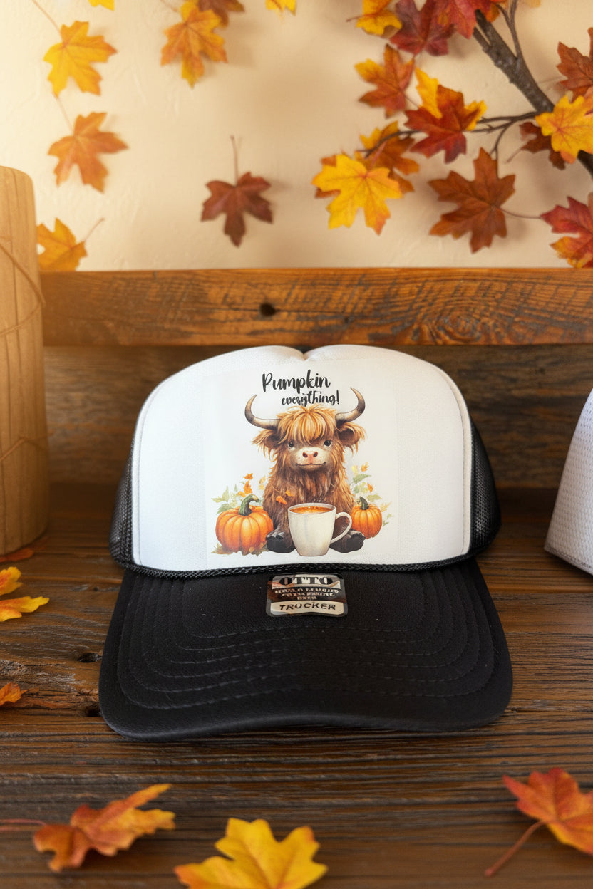 A black and white trucker hat with a custom graphic of a shaggy Highland cow, pumpkins, and a coffee mug, resting on a rustic wooden surface.