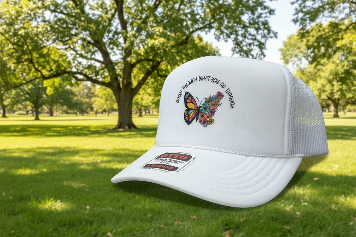 A white trucker hat with a mesh back, featuring a butterfly and floral graphic on the front with the text 'GROW THROUGH WHAT YOU GO THROUGH.' A sewn-on brand patch for Otto Trucker is visible on the brim, and the hat is photographed on a wooden surface.