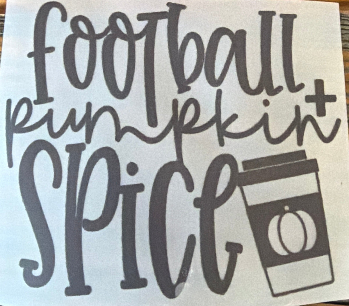 A square white sticker or decal with the phrase 'football pumpkin spice' in dark gray lettering and a small coffee cup graphic, placed on a brown wood-grain tabletop.