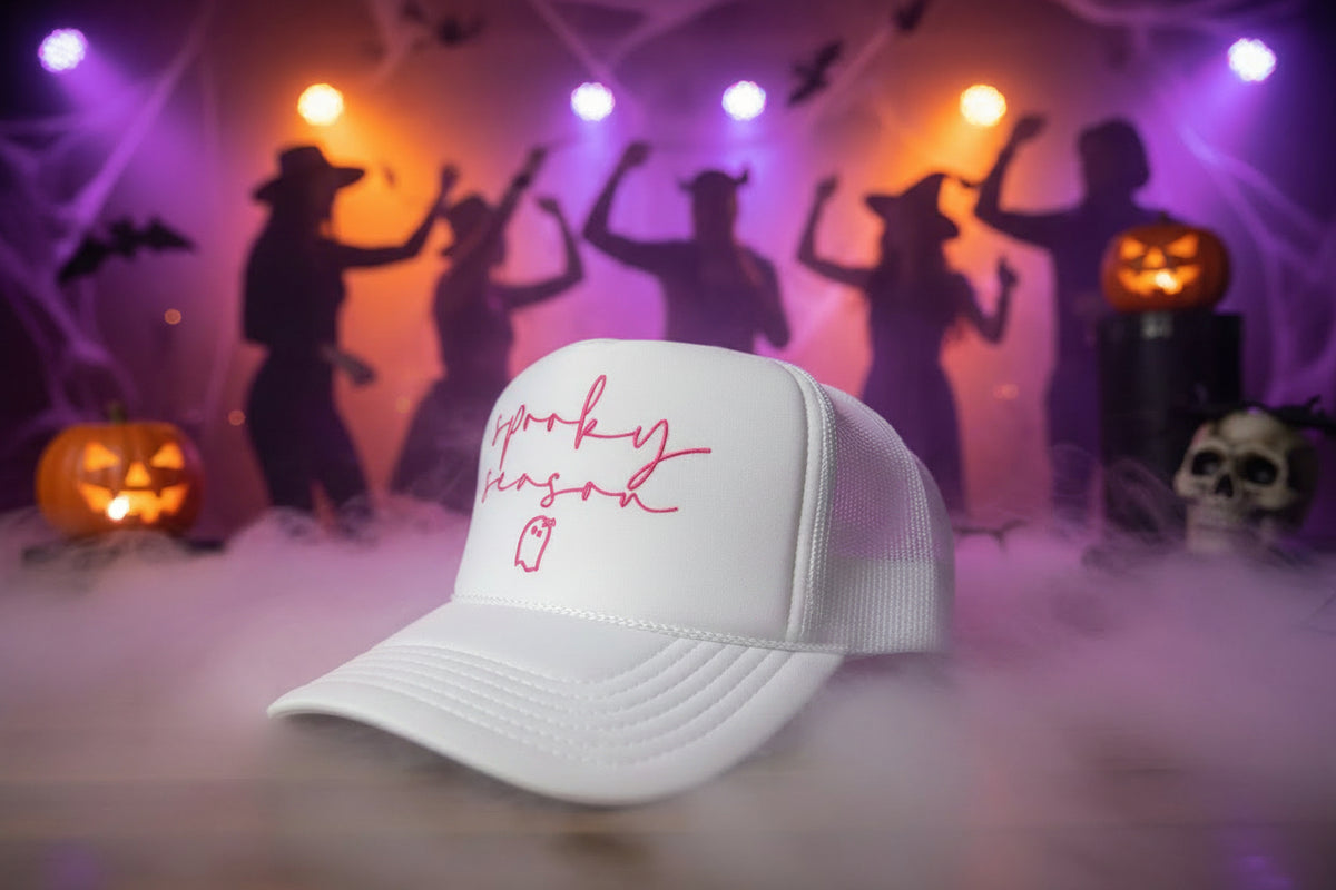 A white trucker hat with pink script text reading 'spooky season' and a small ghost outline on the front, resting on a wooden surface. A visible sticker on the brim reads 'OTTO TRUCKER'.