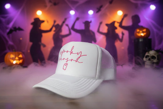 A white trucker hat with pink script text reading 'spooky season' and a small ghost outline on the front, resting on a wooden surface. A visible sticker on the brim reads 'OTTO TRUCKER'.