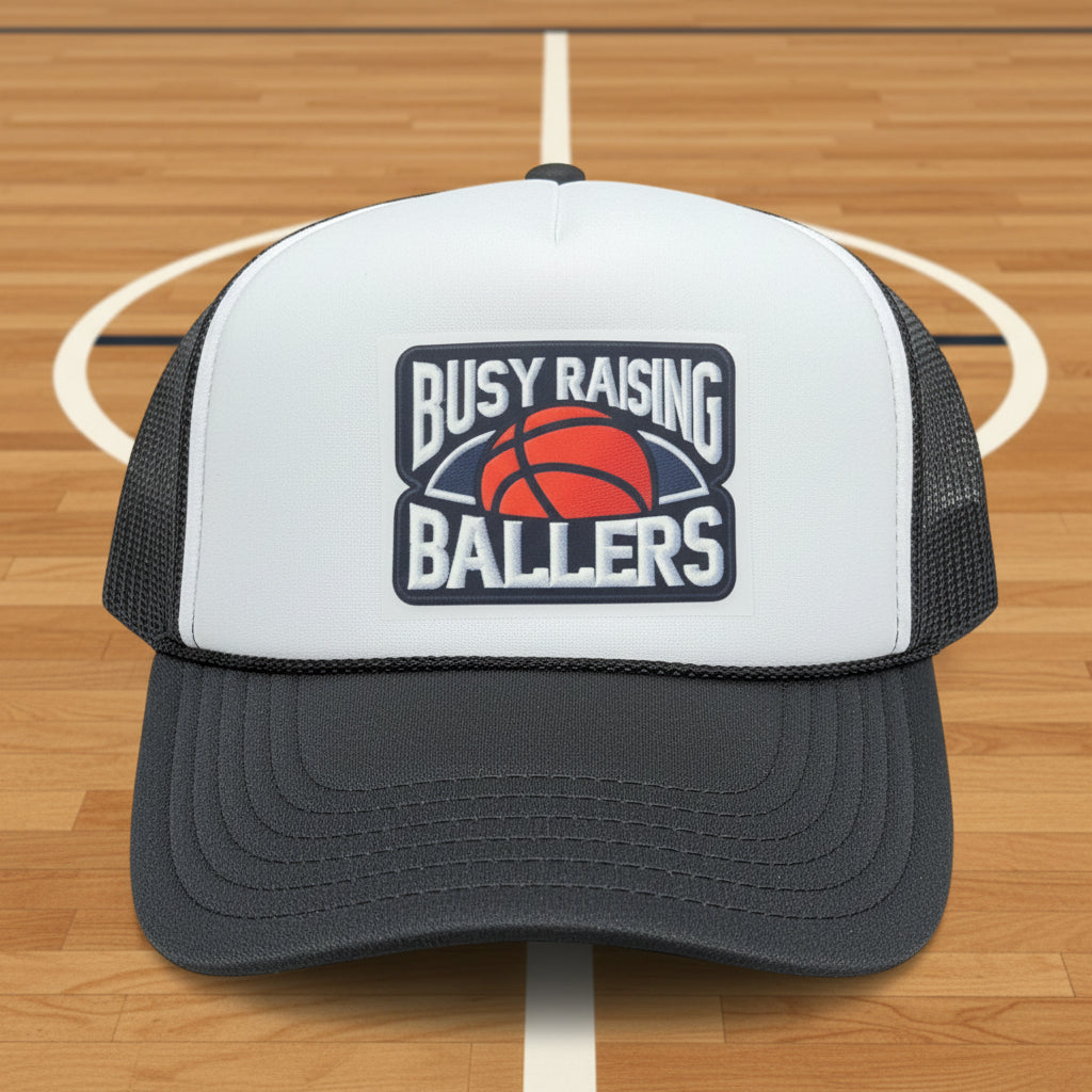 A white and dark gray trucker hat with a 'Busy Raising Ballers' basketball logo on the front.