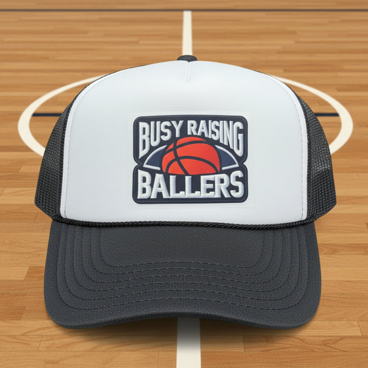 A white and dark gray trucker hat with a 'Busy Raising Ballers' basketball logo on the front.