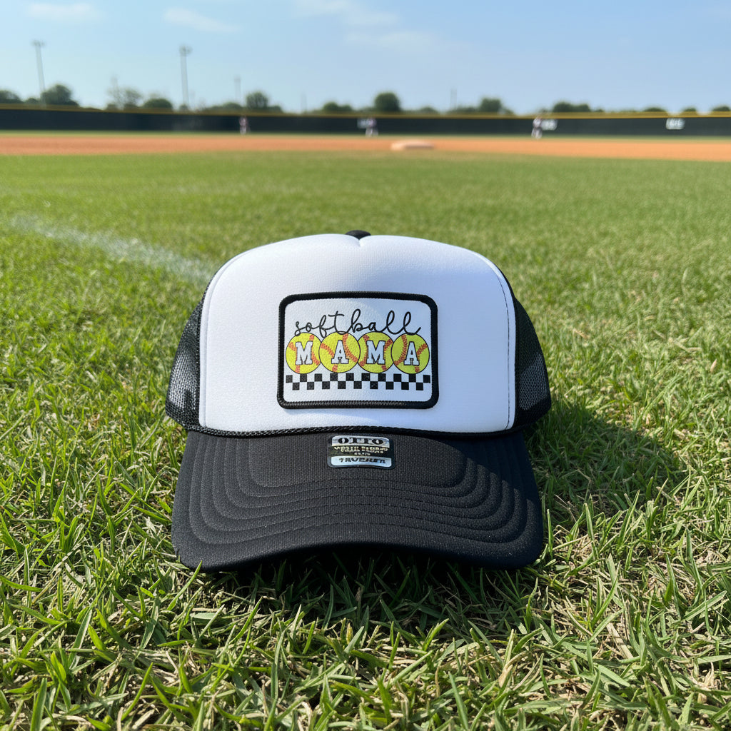 A black and white trucker hat with a front patch reading 'softball MAMA' in yellow and orange patterned circles. 