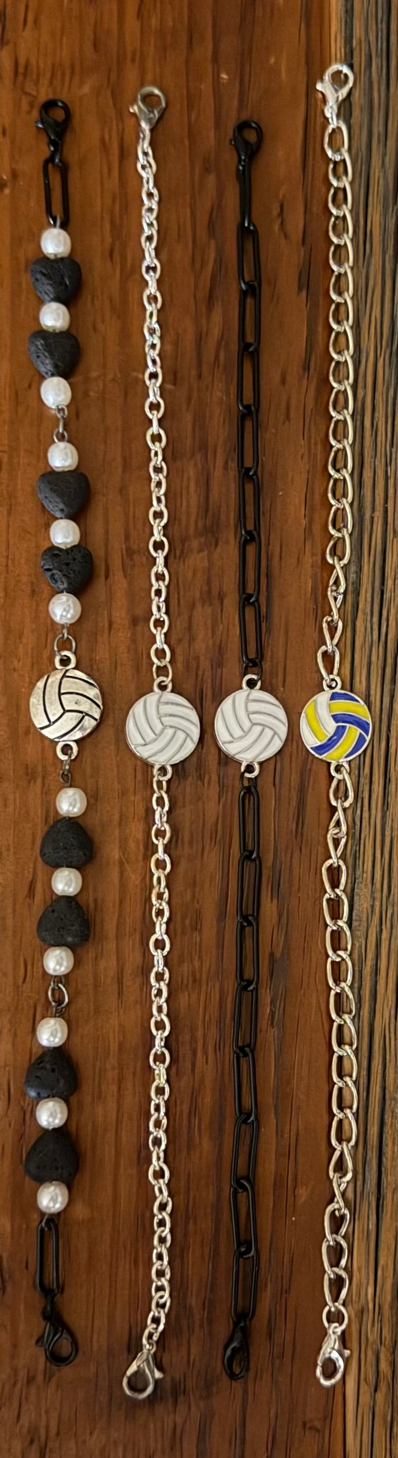 Four beaded and chain bracelets laid flat on a dark wood background. Three bracelets feature black and white beads with silver-toned links, and one bracelet features a yellow and blue volleyball charm on a silver chain.