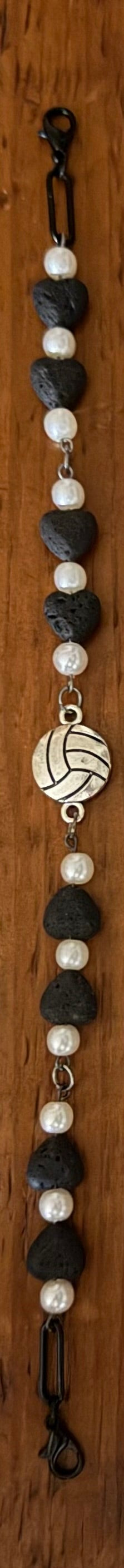Four beaded and chain bracelets laid flat on a dark wood background. Three bracelets feature black and white beads with silver-toned links, and one bracelet features a yellow and blue volleyball charm on a silver chain.