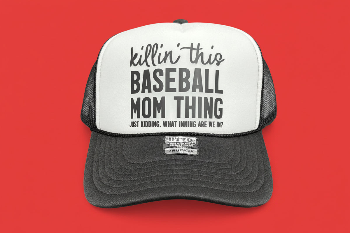 A trucker-style baseball cap with a white front panel and dark gray mesh sides and brim, displayed on a black table against a dark wood-paneled wall. The front reads 'killin' this BASEBALL MOM THING' with smaller text below.
