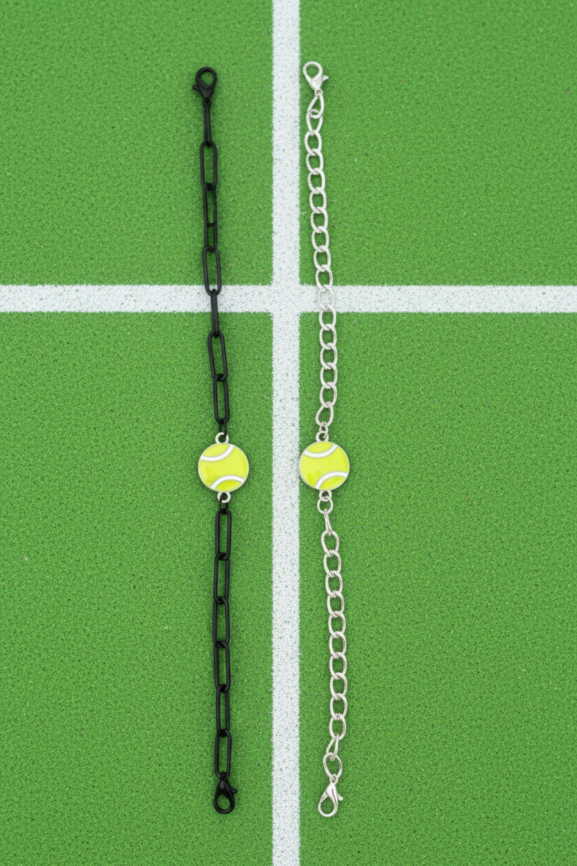 Two tennis-themed chain accessories displayed on a wooden surface: one black chain with two green and white tennis ball charms, and one silver chain with two matching tennis ball charms.