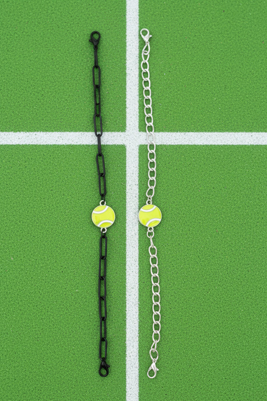Two tennis-themed chain accessories displayed on a wooden surface: one black chain with two green and white tennis ball charms, and one silver chain with two matching tennis ball charms.