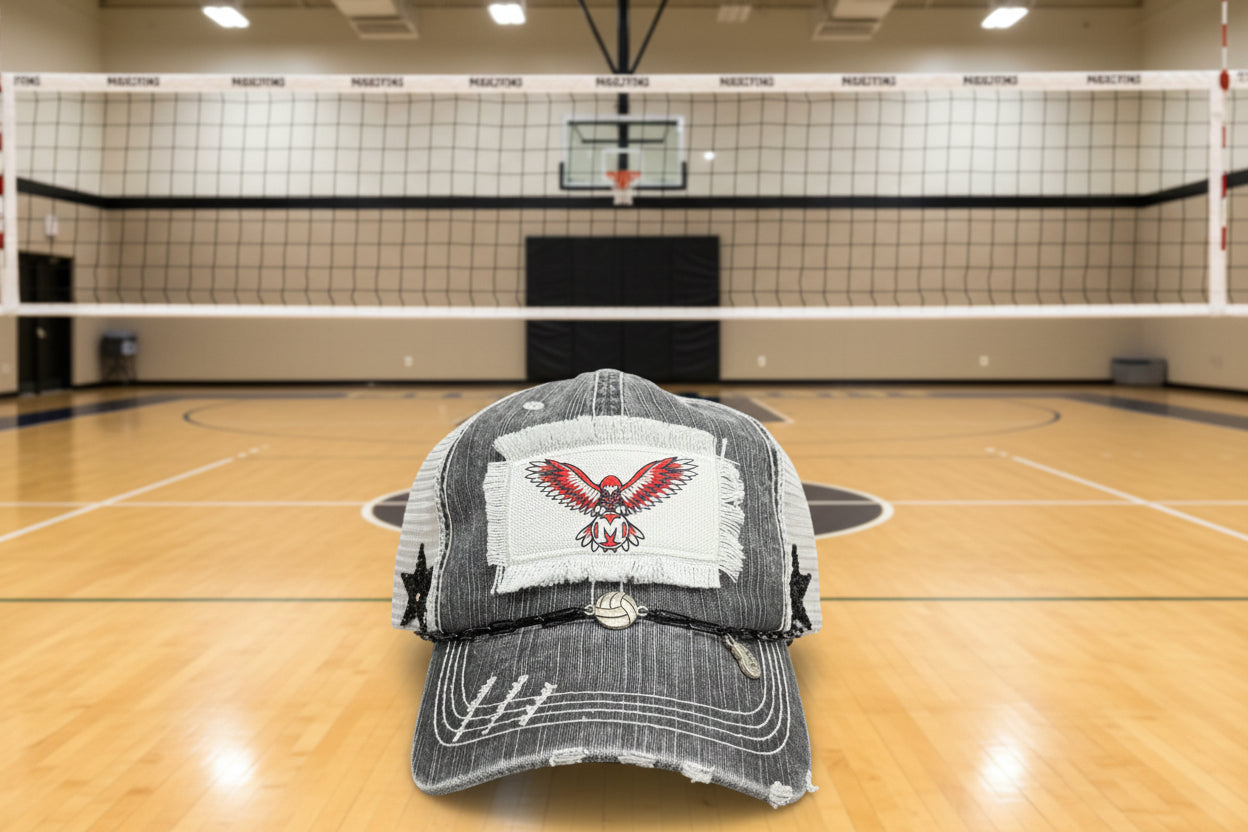 A distressed gray and white trucker cap with a white patch featuring a red and white eagle with the letter 'M'. A black paperclip hat chain with a volleyball and a heart mom charm rests across the brim.
