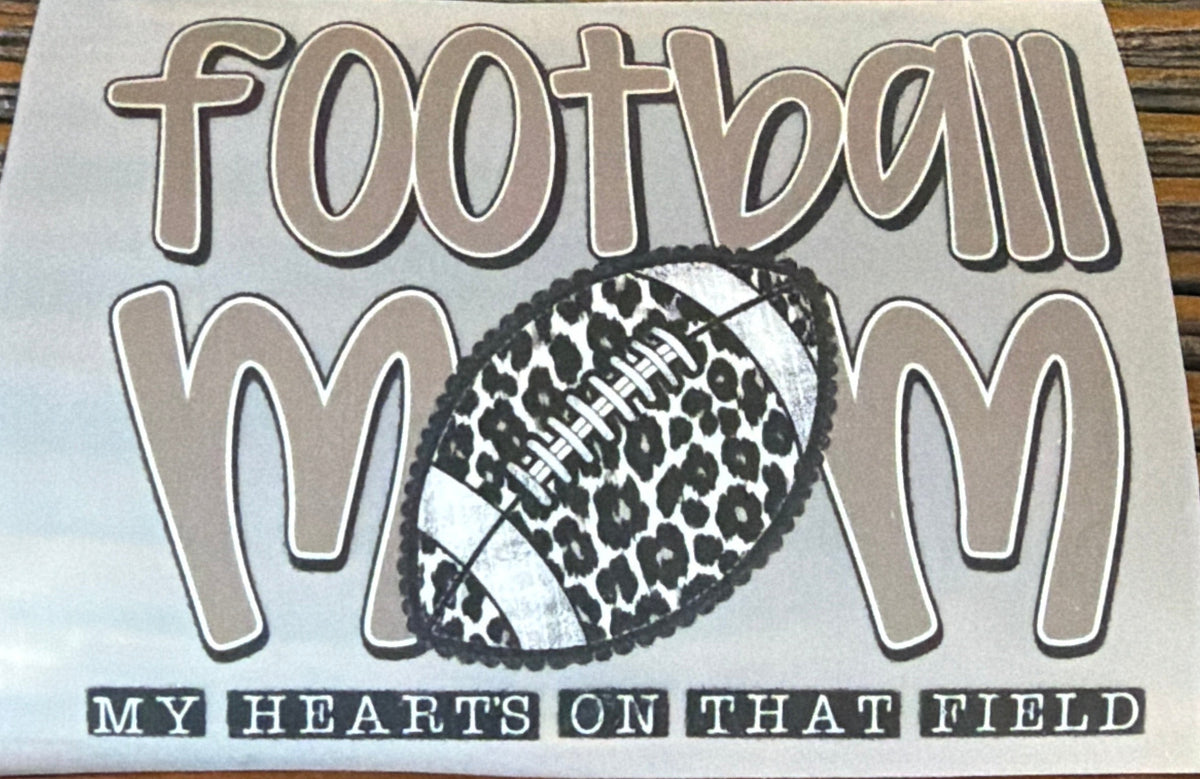 A die-cut vinyl sticker with the text 'football mom' and 'MY HEARTS ON THAT FIELD' in black and gray, featuring a leopard-print and white football graphic in the center, displayed on a dark wood surface.