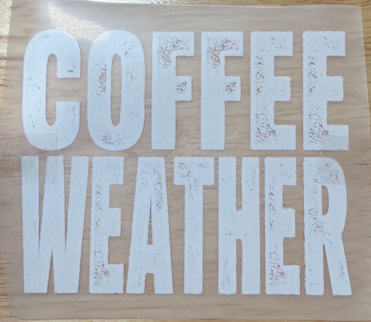 A vinyl decal with the words 'COFFEE WEATHER' in distressed white block letters on a clear backing sheet, placed on a light wood tabletop. A person's legs and feet are visible at the bottom edge of the frame.