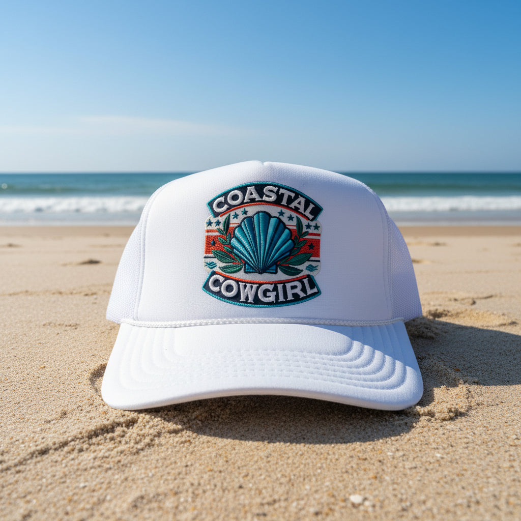 A white trucker hat with a mesh back, featuring a large embroidered patch on the front that reads 'COASTAL COWGIRL' around a blue shell motif. A white paper label on the brim reads 'OTTO World Famous Foam Front OS38 Trucker'.