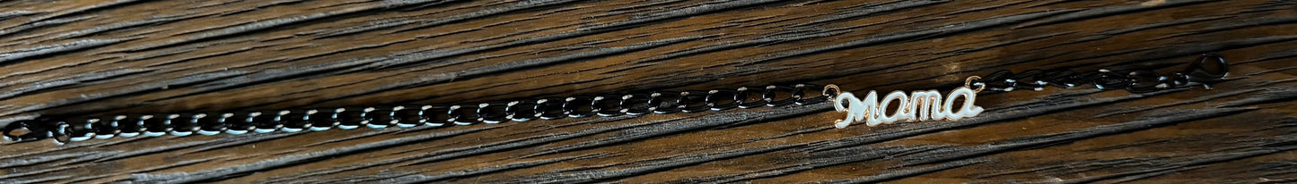 A dark metal chain bracelet with a cursive 'mama' nameplate pendant, displayed on a dark wood background.