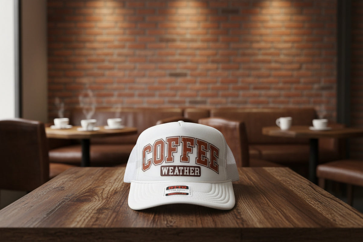 A white trucker hat with 'COFFEE WEATHER' printed on the front in brown and white, resting on a wooden table.