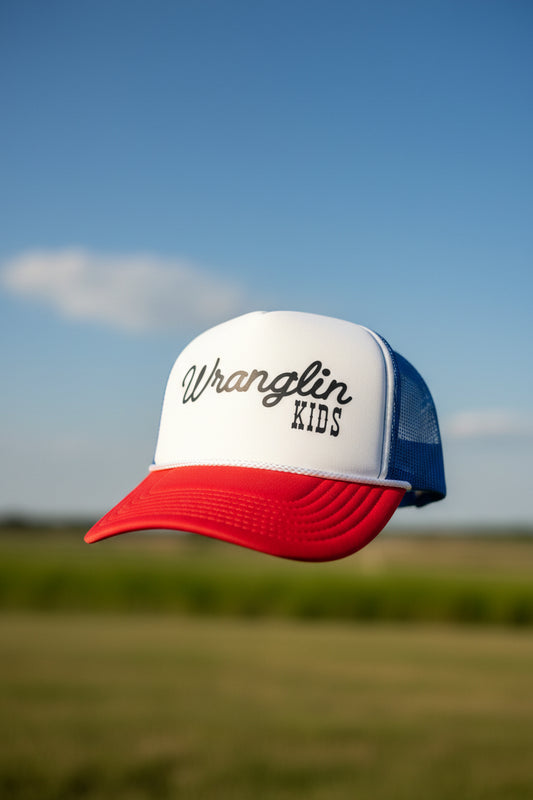 A red, white, and blue trucker hat with 'Wrangler KIDS' DTF print on the front, displayed on a black table against a dark wood panel background.
