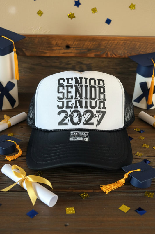 A black and white trucker hat with 'SENIOR SENIOR class of 2027' printed on the front, resting on a rustic wooden surface.