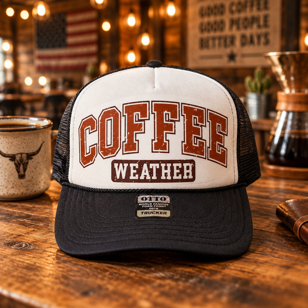 A black and white trucker hat with 'COFFEE WEATHER' embroidered on the front, displayed on a wooden table with other hats in the background.
