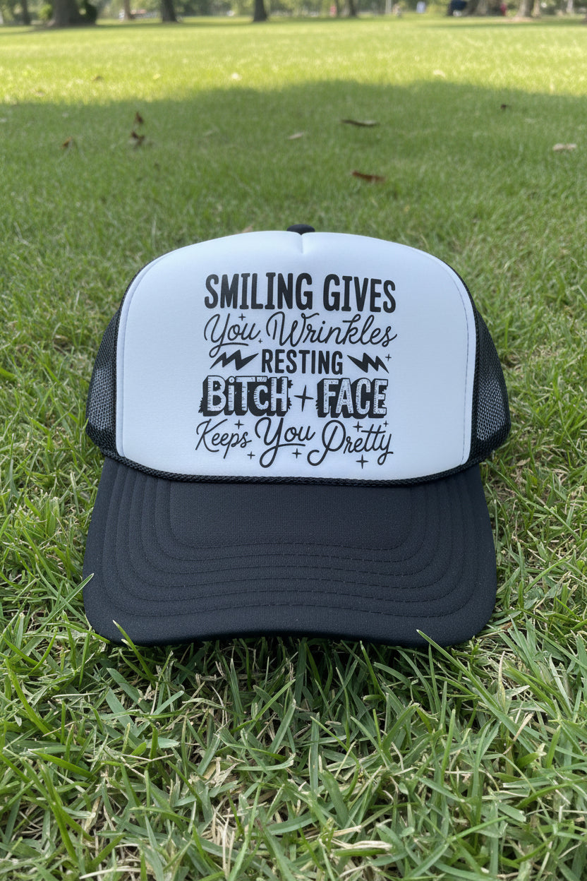 A black and white trucker hat with a humorous quote printed on the front panel. The hat features a white front panel with black text that reads 'SMILING GIVES You Wrinkles RESTING BITCH + FACE Keeps You Pretty' and a black brim.