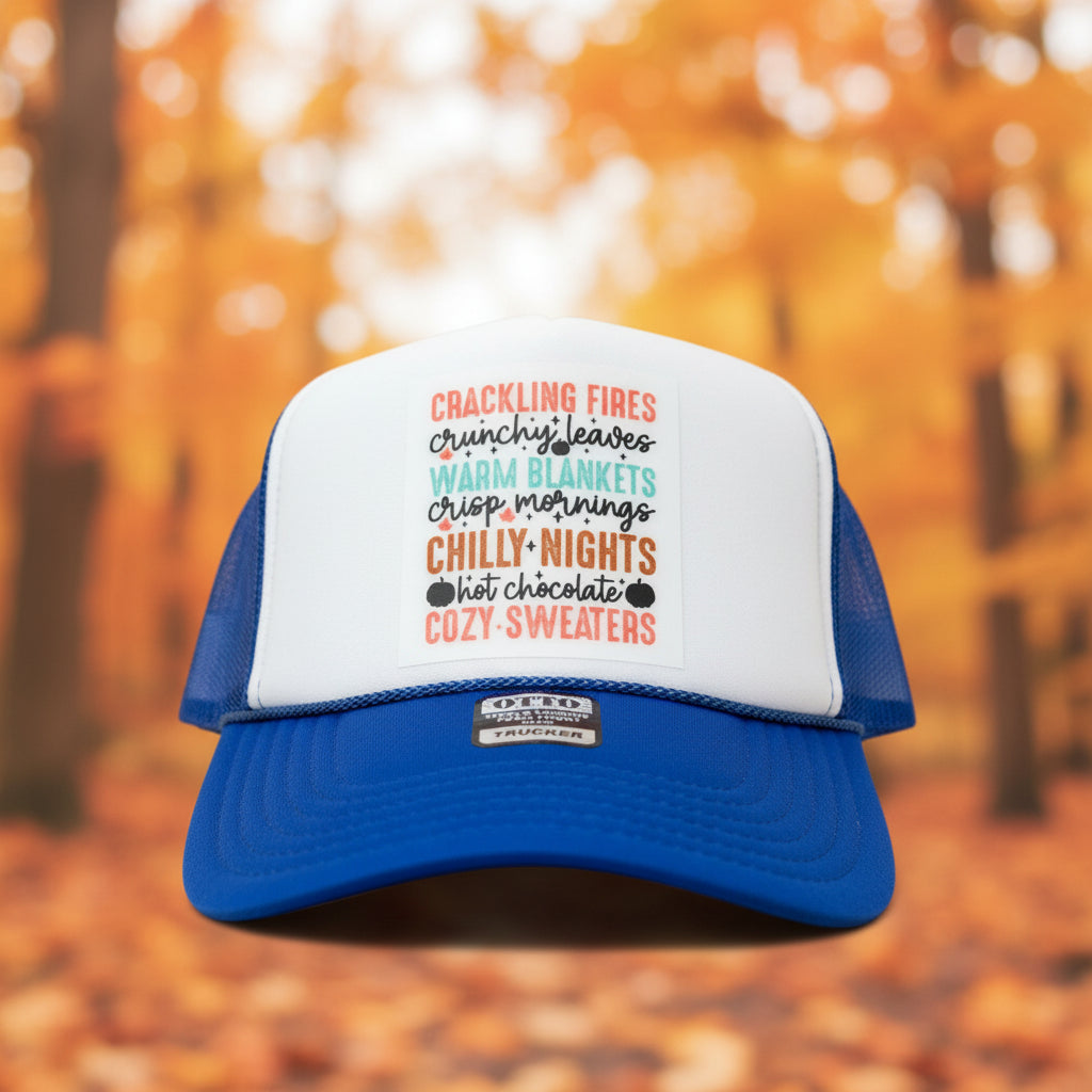 A blue and white trucker hat with a custom printed list of cozy things on the front panel, resting on a rustic wooden surface.