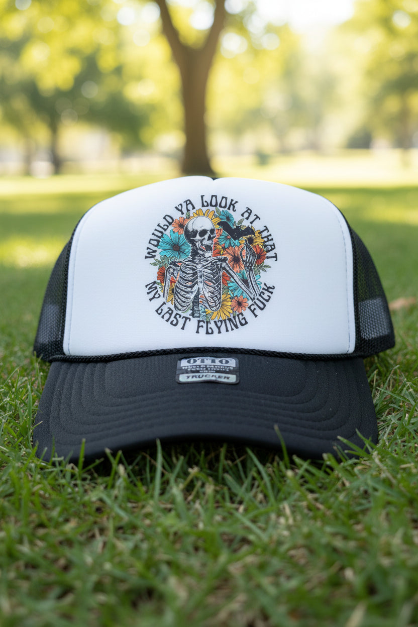 A black and white trucker hat featuring a graphic of a skeleton surrounded by flowers and birds, with the text 'WOULD YA LOOK AT THAT MY LAST FLYING FUCK'. The hat has a white front panel, black mesh back, and a black brim with a visible 'OTTO' brand sticker.