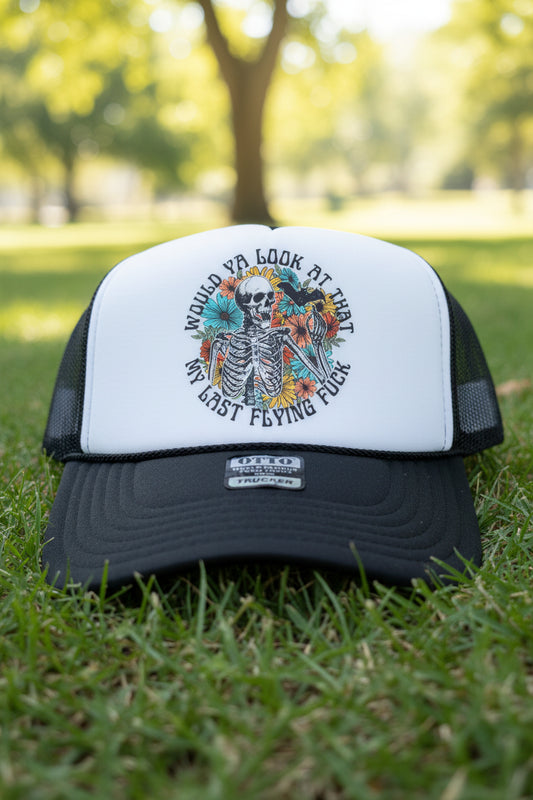 A black and white trucker hat featuring a graphic of a skeleton surrounded by flowers and birds, with the text 'WOULD YA LOOK AT THAT MY LAST FLYING FUCK'. The hat has a white front panel, black mesh back, and a black brim with a visible 'OTTO' brand sticker.