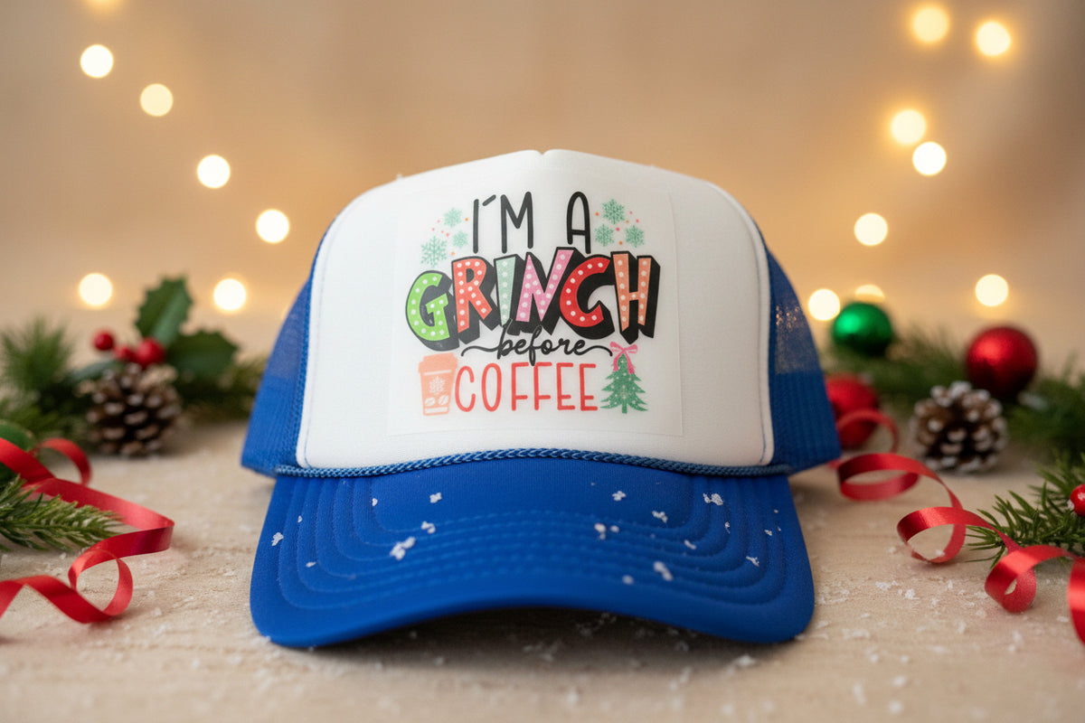 A blue and white trucker hat with a festive graphic that reads 'I'M A GRINCH before COFFEE' in colorful lettering, resting on a rustic wooden surface.