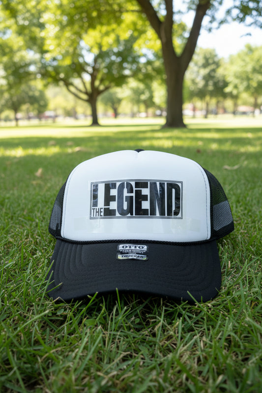 A black and white trucker hat with 'THE LEGEND' printed on the front panel, resting on a light wood surface.