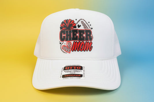 A white trucker hat with a 'Cheer Mom' graphic on the front, displayed on a dark wooden surface with a dark wood background.